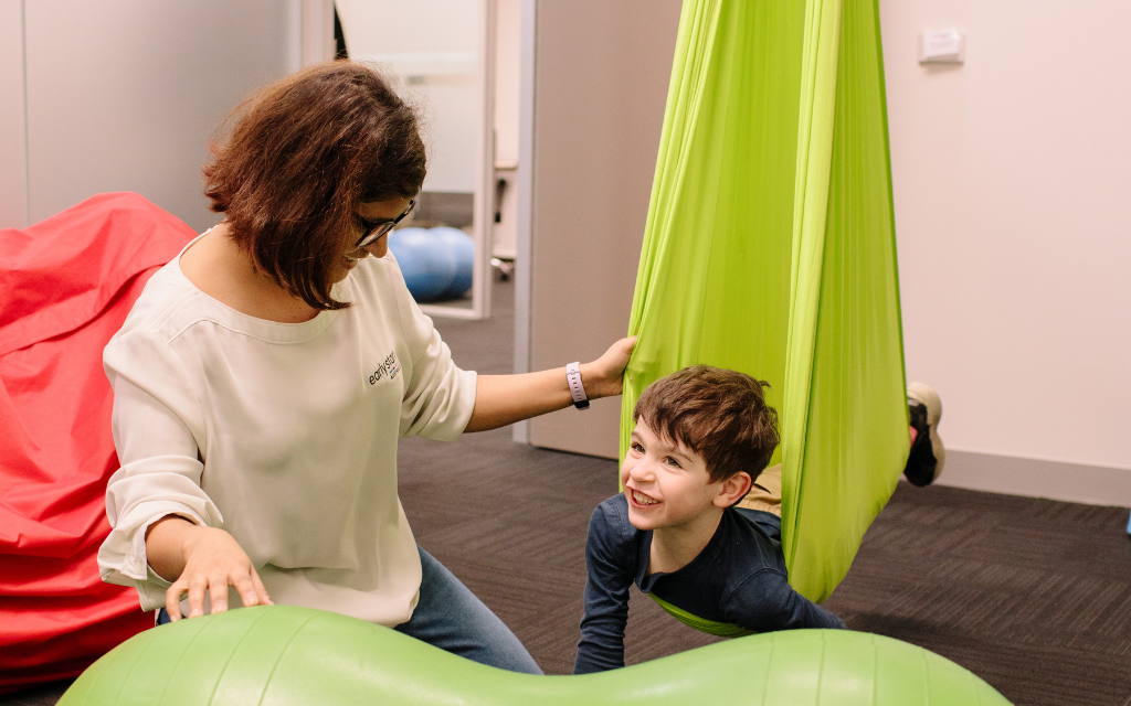 Young boy swinging on sensory gym swing smiling at his female occupational therapist. 